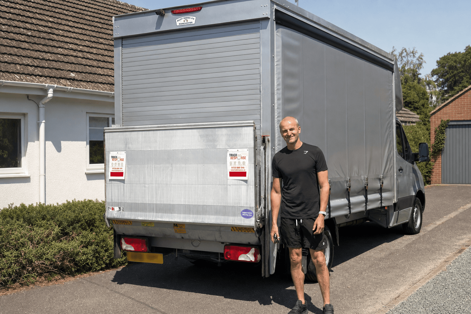 Aidan standing at the rear of the curtainside Luton outside a house, next to the roller shutter and the tail lift platform folded up against it.
